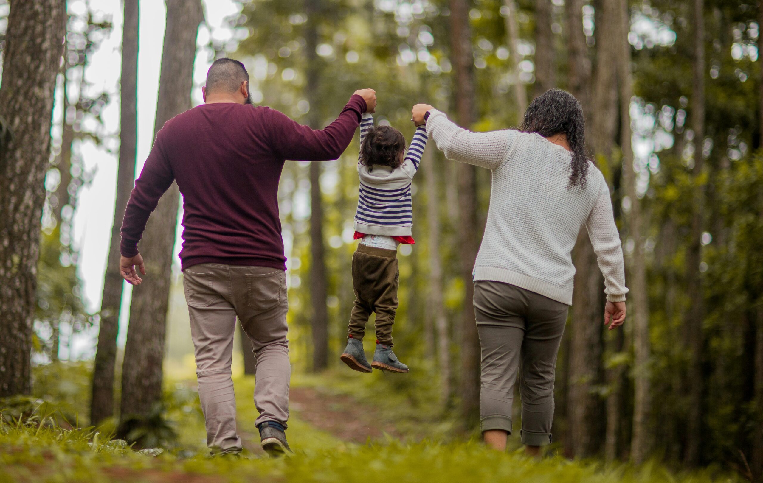 A family of three walks hand-in-hand through a forest, joyfully swinging a child between them.